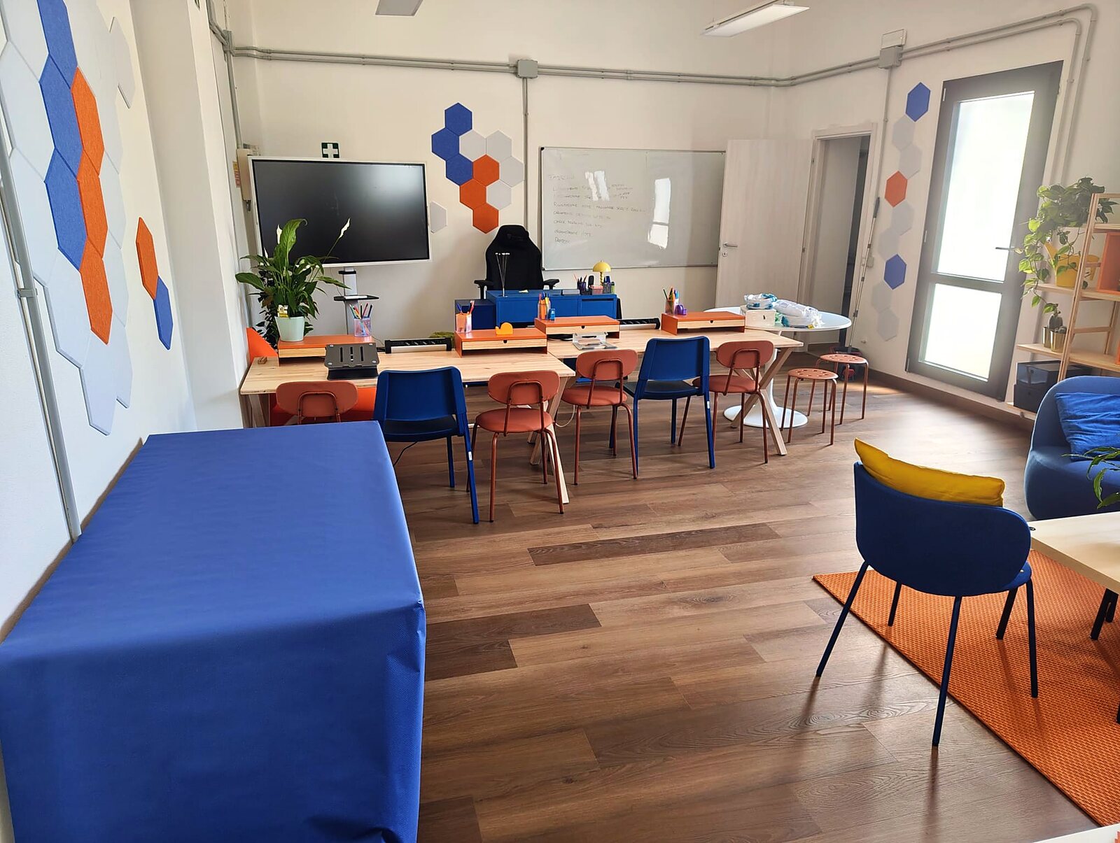 L'aula dello Spazio Binatomy: tavoli in legno, schermo grande, lavagna e pannelli esagonali blu e arancio